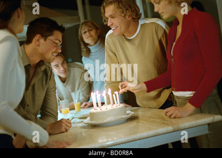 Woman lighting candles on birthday cake, friends standing nearby smiling, watching Stock Photo