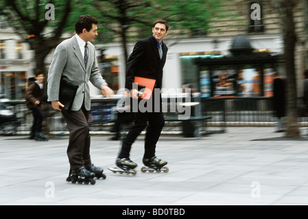 Men in business attire inline skating together along sidewalk carrying briefcase, folders under arm Stock Photo