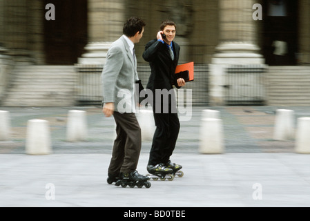 Men in business attire inline skating together along sidewalk, one phoning Stock Photo