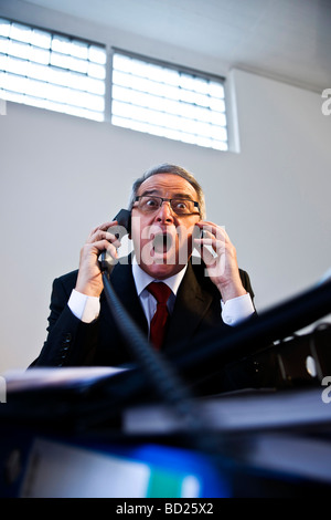 Manager with glasses and suit sitting in front of a mountain of folders, angrily speaking on two telephone lines Stock Photo