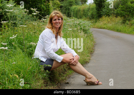 Mature woman wearing a white shirt and skirt sitting on the roadside Stock Photo