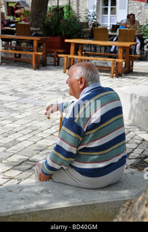 elderly retired old man sitting relaxing looking observing resting on walking stick in the shade on bright sunny day in France Stock Photo