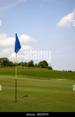 Golf green with hole, flagstick, flag, bunker, blue sky, and trees in the background. Stock Photo