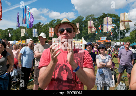 Close up portrait of a WOMAD music festival man with trilby and sunglasses clapping with a festival crowd and flags behind him Stock Photo