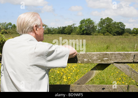 Elderly man leaning on five barred gate looking over a field of flowers to a countryside view Stock Photo