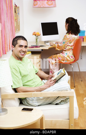 Portrait of a mature man reading a book and listening to an mp3 player with a mid adult woman using a laptop in the background Stock Photo