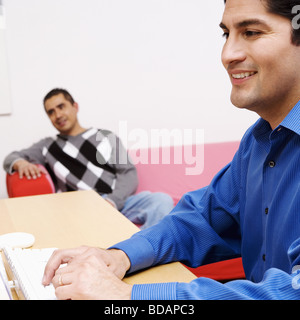 Side profile of a businessman working on a desktop PC with a mature man sitting on a couch in the background Stock Photo