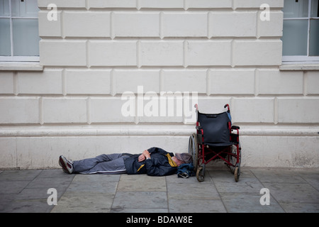 A homeless man lays next to his wheel chair sleeping perhaps passed out on the pavement, London Stock Photo