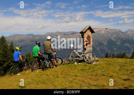 Three mountain bikers stop to admire the view next to a religious shrine in the mountains high above Sauze D'oulx, Italy Stock Photo