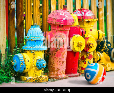 Colorful old fire hydrants painted in bright colors, sitting on sidewalk against wood fence and child's rubber ball. Stock Photo