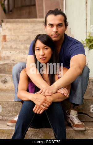 Male and female couple sitting on concrete steps, his arms are around her.  Looking at camera.  Woman is Filipino. Stock Photo