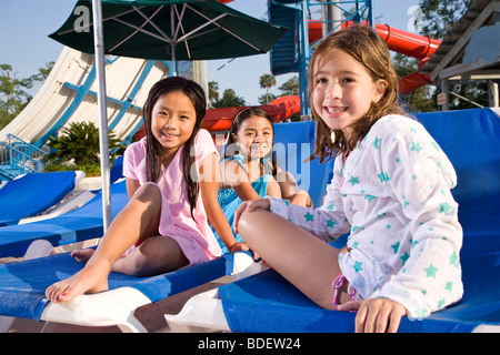 Multi-ethnic girls sitting on deck chairs at water park in summer Stock Photo