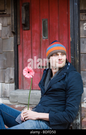 Young stylish man holding flower sitting on cobblestone sidewalk Stock Photo