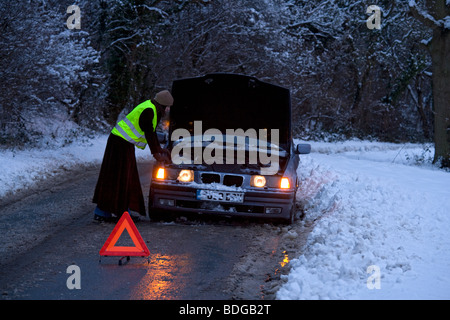 Women on her own broken down in the snow with warning triangle, stranded trying to get it fixed wearing fluorescent jacket Stock Photo