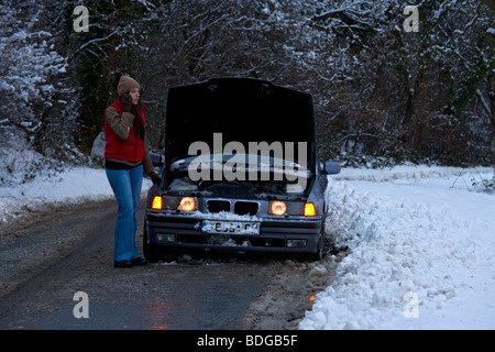Women on her own broken down in the snow, stranded trying to get it fixed on mobile phone ringing rescue service at night. Stock Photo