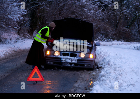 Women on her own broken down in the snow at night , with reflective jacket, warning triangle and torch trying to get it fixed. Stock Photo