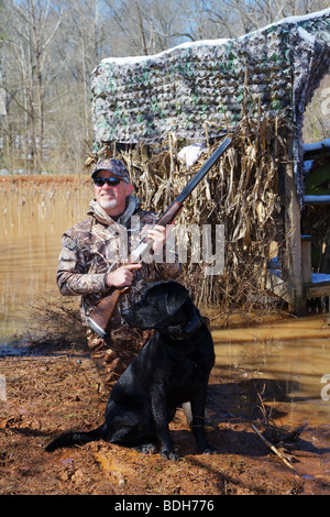 DUCK HUNTER IN CAMO AND BLACK LAB LABRADOR ON SHORE WAITING FOR WATERFOWL MALLARDS BROWNING CITORI SHOTGUN SNOW ON GROUND BLIND Stock Photo