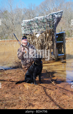 DUCK HUNTER IN CAMO AND BLACK LAB LABRADOR ON SHORE WAITING FOR WATERFOWL MALLARDS BROWNING CITORI SHOTGUN SNOW ON GROUND BLIND Stock Photo