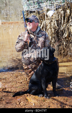 DUCK HUNTER IN CAMO AND BLACK LAB LABRADOR ON SHORE WAITING FOR WATERFOWL MALLARDS BROWNING CITORI SHOTGUN SNOW ON GROUND BLIND Stock Photo