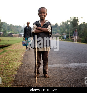 A boy with a walking stick poses on the edge of a paved road with villagers walking the road behind him. Stock Photo