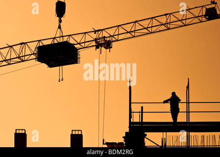 construction site in the morning, silhouetted worker on construction frame conducting crane with heavy load Stock Photo