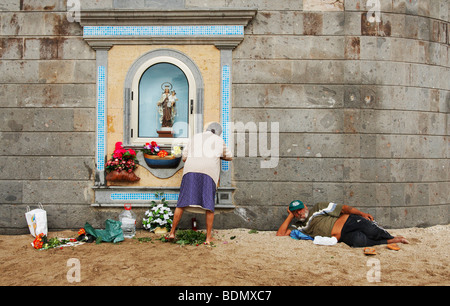 Old Spanish woman changing flowers at shrine on beach while man watches. Could be used for gender pay gap, equal pay... Stock Photo