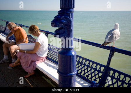 A gull waits for scraps while a couple eat their food on the pier at Eastbourne, England. Stock Photo