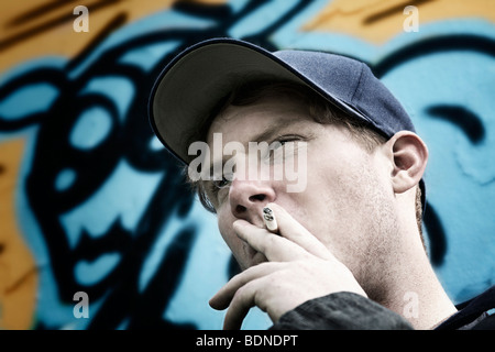 Portrait of a young man smoking a cigarette, wearing a cap, in front of a graffiti-sprayed wall Stock Photo
