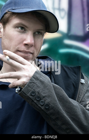 Portrait of a young man smoking a cigarette, wearing a cap, in front of a graffiti-sprayed wall Stock Photo