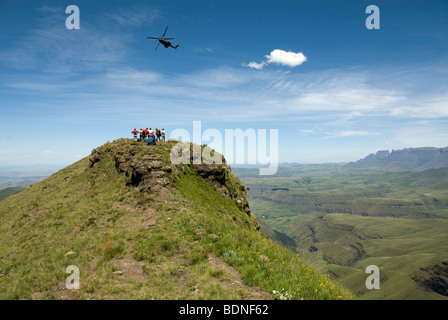Oryx helicopter passing hikers on rocky outcrop, Cathkin Peak and Little Berg in distance, Cathedral Peak Range Stock Photo