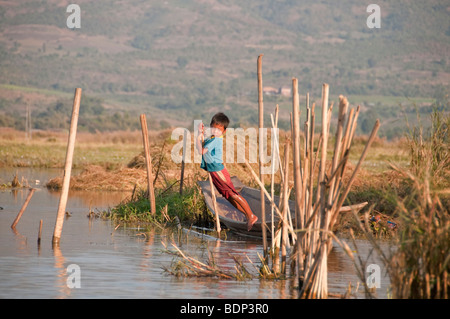 Young Shan boy taking a break from practicing one-legged rowing to lean on a bamboo stake from their family's floating island in Stock Photo