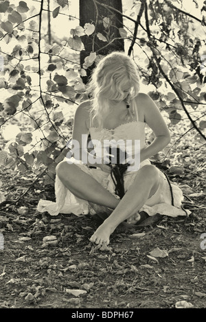 A young woman sitting cross legged wearing a white summer dress holding a red heart sitting beneath a tree in b/w