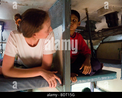 Western European passenger resting on a sleeper bed, & talking to a local boy passenger in a sleeper compartment. Indian train. Stock Photo
