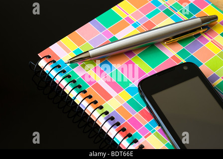 Black cell phone with colorful notebook and silver pen isolated on black background Stock Photo