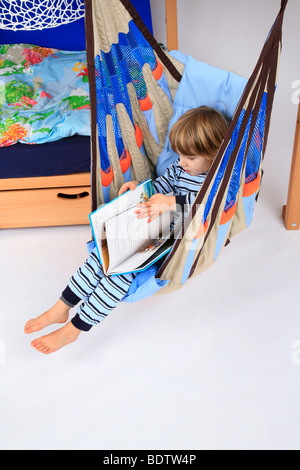 little boy in pyjama reading a children's book in a swing of a Billi-Bolli loft bed Stock Photo