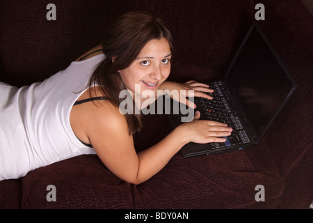 Pretty brunette teenage girl laying on the sofa and typing on laptop Stock Photo