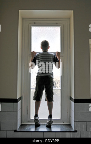 Lonely nine-year-old boy in the hallway, looking through the opal glass window to the backyard, Germany, Europe Stock Photo