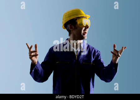 Workman wearing a boiler suit, a yellow hard hat and protective goggles is gesticulating Stock Photo