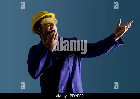 Workman wearing a boiler suit, a yellow hard hat and protective goggles looks frightened Stock Photo