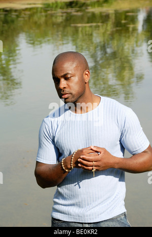 Young black man holding rosary looking thoughtful by pond Stock Photo