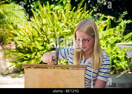 Tween tweens young person people  girl 11-13 year old repairing cutting board by hammering nail  into wooden cutting board eyeglasses  MR Myrleen Pearson Stock Photo