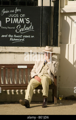 Elderly Man In A Suit And Fedora Relaxing In The Sun With A Glass Of Wine On A Bench Below A Sign For Pimms Stock Photo
