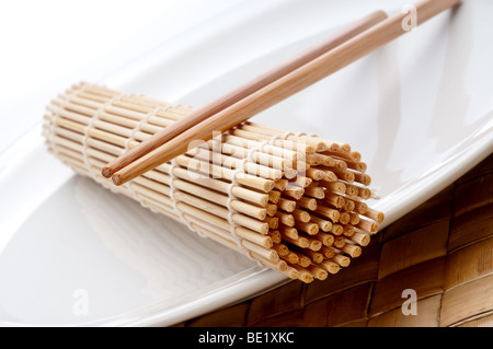 a tilted horizontal macro of a pair of chopsticks and a sushi rolling mat on a white plate Stock Photo