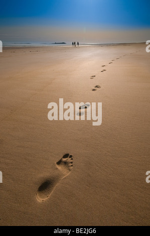 Footsteps in the sand on a golden beach with a group of people in the distance Stock Photo