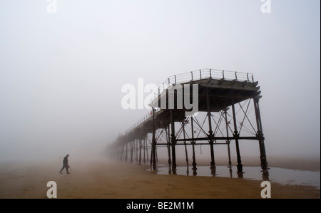 A metal detector searches the beach and sand on a misty day under the legs of a victiorian pier on the east coast of England. Stock Photo