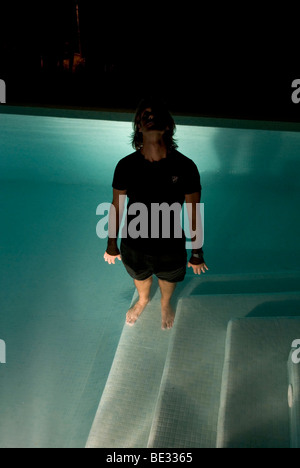a strange eerie photo of dressed man standing up to his waist in swimming pool at night illuminated by pool lights Stock Photo