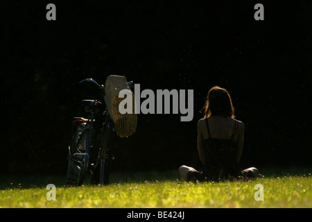 Young adult woman sitting on lawn grass meadow with bicycle on sunny summer day with a lot of flies around on black background Stock Photo