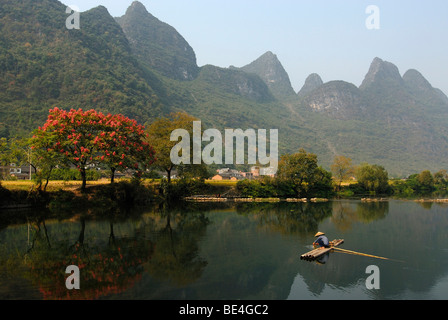 Chinese fisherman with a straw hat on a bamboo raft fishing near Yangshuo in the Yulong River in front of flowering trees and l Stock Photo