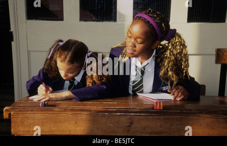 Two little girls in a classroom.  One girl is taking a sneaky peek her friend's work.
