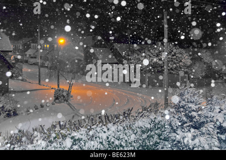 Night snowing flakes camera flash orange lamp post light onto snow covered road in residential landscape manipulated mainly black & white England UK Stock Photo